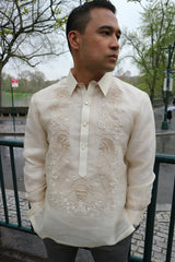 Ricky wears a hand embroidered piña silk Barong Tagalog and grey pants. He wears a chamisa de chino underneath his barong. He stands with his hands in his pockets and he looks to his left. Ricky stands in front of a green gate, there is cobblestone sidewalk, a street and Central Park is behind him. The skies are grey and gloomy