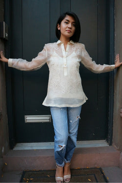 Robin stands in front of a dark green door in her hand embroidered piña silk Barong Tagalog, white tank top underneath her barong, blue jeans and tan open toe shoes. She stands with her hands reaching and touching the sides of the doorway