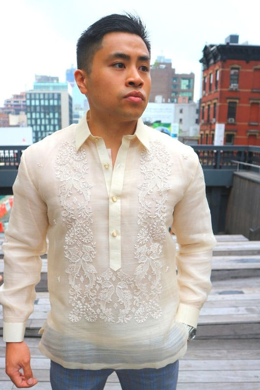 Rodrigo stands in his hand embroidered piña silk Barong Tagalog, chamisa de chino, and grey and blue plaid slacks. He stands with his left hand in his pocket, his right hand hanging to his side, and looking to his left. Rodrigo stands at the High Line Park in New York City with stairs, a railing and buildings behind him.