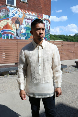 Ronnie stands in his hand embroidered piña silk Barong Tagalog, a chamisa de chino underneath his barong and black pants in front of stone benches, a fence, a building with a mural, trees and the sky in the background