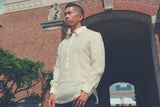 Upward angled photo of Ronnie in his hand embroidered piña silk RonRiz Barong Tagalog. Ronnie wears a chamisa de chino underneath his barong and black pants. He stands at the entrance of the Univresity of Cincinnati. There is a tree, brick building and tunnel hall leading to another building behind him