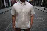 Product photo of the short sleeve hand embroidered jusi Ryan Barong Tagalog. Ryan wears a chamisa de chino underneath his barong, brown slacks and a silver watch on his left wrist. Ryan is standing on a cobblestone road with downtown NYC buildings around him. There are people walking on the sidewalks on both sides of Ryan