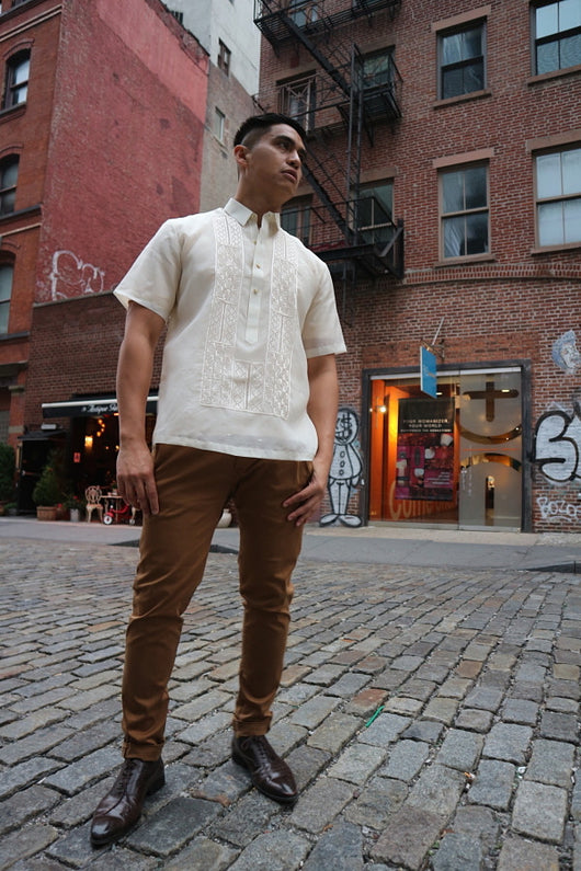 Upward photo of Ryan standing on a cobblestone road with downtown NYC buildings behind him. Ryan wears a short sleeve hand embroidered jusi Barong Tagalog, a chamisa de chino underneath his barong, brown slacks and dark brown shoes