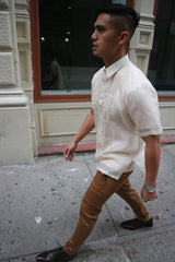 Side view of Ryan walking on the sidewalk with a building with windows to his right. Ryan wears a short sleeve hand embroidered jusi Barong Tagalog, a chamisa de chino underneath his barong, brown slacks, dark brown shoes and a silver watch on his left wrist