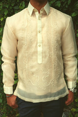 Product photo of the hand embroidered cocoon Ryan Barong Tagalog. Ryan wears a chamisa de chino underneath his barong, blue navy slacks, a silver watch on his left wrist and a bracelet on his right wrist. Ryan stands in front of a green leaf covered wall with his thumbs hanging onto his pants pockets  