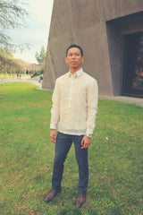 Ryan stands on a grass lawn with a cement structure behind him. There are paved walkways, trees and people walking and sitting in the distant background. Ryan wears a hand embroidered cocoon Barong Tagalog, a chamisa de chino underneath his barong, navy slacks, brown shoes and a silver watch on his left wrist. Ryan stands straight at an angle as he looks to his left