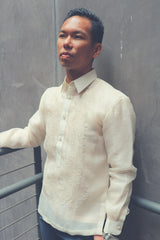 Photo of Ryan from the hips up. Ryan standing at an angle looking straight ahead with his back to a grey wall, his right arm resting on grey handrails and his left hand in his pocket. Ryan wears a hand embroidered cocoon Barong Tagalog. Ryan wears a chamisa de chino underneath his barong, navy slacks and a silver wrist watch on his left wrist.