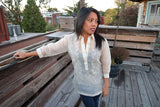 Sarahlynn stands on a wooden rooftop deck with a wooden wall next to her and behind her. There are other buildings, rooftops, trees and the sky in the background. Sarahlynn wears a hand embroidered piña silk Barong Tagalog, black tank top underneath her barong and dark blue jeans. She stands with her right hand leaning and resting on the wooden wall to her right. She is looking to her left with her left hand resting at her side