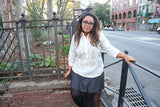 Stephanie stands in her hand embroidered jusi Barong Tagalog, a black 3/4 sleeve shirt underneather her barong, grey skirt and black leggings. She stands with her back leaning on a park gate behind her and on her left side with her head tilted slightly to her left, her left hand rests on the black rail next to her and her right hand resting on the gate behind her. There is a tree and plant area of a park behind her and a street with parked cars and buildings are to her left
