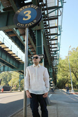 Upward photo of Steven leaning next to a street sign that says Entrance 3 Citi Field. Steven stands on the sidewalk on Roosevelt Avenue under the elevated tracks of the 7 train in Flushing. Steven wears a hand embroidered piña silk Barong Tagalog, a chamisa de chino underneath his barong, dark jeans and black sunglasses. He stands with his hands hanging off his pockets. There are streets, a pedestrian, trees, a street light, other street signs, and the blue sky in the background