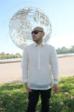 Steven stands in Flushing Meadow Park in front of the Unisphere. Steven wears a hand embroidered piña silk Barong Tagalog, a chamisa de chino underneath his barong, dark jeans and black sunglasses. He stands on the grass and there are trees and the blue sky in the background