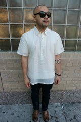 Steve stands in his short sleeve hand embroidered jusi Barong Tagalog, a chamisa de chino underneath his barong, dark jeans, black sunglasses, brown shoes and black bracelet on his left wrist. He stands in front of a brick wall with square glass windows on the top half of the wall above the bricks
