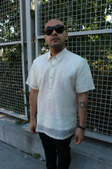 Steve stands in his short sleeve hand embroidered jusi Barong Tagalog, a chamisa de chino underneath his barong, dark jeans, black sunglasses and black bracelet on his left wrist. He stands in front of a grid gate with trees behind it