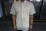 Product photo of the short sleeve hand embroidered jusi Steve Barong Tagalog. Steve wears a chamisa de chino underneath his barong, dark jeans, and black bracelet on his left wrist. He stands in front of a storefront with windows with the street, cars and people reflected in the glass