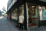 Angled photo of Steve in his short sleeve hand embroidered jusi Barong Tagalog, a chamisa de chino underneath his barong, dark jeans, brown shoes, black sunglasses and black bracelet on his left wrist. He stands in front of a Filipino grocery store in Little Manila, Queens, NYC. You can see down the sidewalk on the left side of the picture