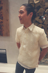 Angled photo of Tiger standing with his hands behind his back and a smirk on his face in front of a modern fireplace wall in his hand embroidered cocoon Barong Tagalog, a chamisa de chino underneath his barong and dark jeans.