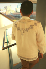 Tonilyn stands with back to camera. Tonilyn is next to a metal and glass pane handrail. There is a window and white and grey wall in the background. Out the window you can see neighboring buildings. We can see the back embroidery on the jusi Tonilyn Barong Tagalog. Tonilyn wears a chamisa de chino underneath the barong, and light brown khaki pants 