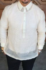 Product photo of the hand embroidered piña silk Zar Barong Tagalog. Zar wears a chamisa de chino underneath his barong, beaded bracelet on right wrist and watch on left wrist, and black pants. Zar stands in front of brown wooden doors 
