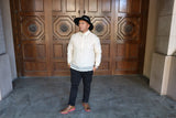 Full body photo of Zar standing in front of brown church doors with hands in his pockets, right foot positioned forward and to right, and looking towards his right. Zar wears a hand embroidered piña silk Barong Tagalog, chamisa de chino underneath his barong, black hat with black bandana tied around the hat, beaded bracelet on right wrist and watch on left wrist, black pants and brown shoes with pink soles. 