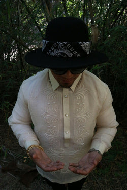 Photo of top half of Zar standing looking down at his palms facing up held in front of his stomach. Zar wears a hand embroidered piña silk Barong Tagalog, chamisa de chino underneath his barong, black sunglasses and black hat with black bandana tied around the hat, beaded bracelet on right wrist and watch on left wrist. There are tree branches and leaves in the dark background
