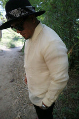 Picture of side view of Zar standing with head tilted down with hands in pockets on a dirt path with trees and branches in the background. Zar wears a hand embroidered piña silk Barong Tagalog, chamisa de chino underneath his barong, black sunglasses and black hat with black bandana tied around the hat, watch on left wrist, and black pants. 