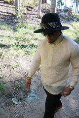 Zar walks on a dirt path next to a hill with grass and palm trees growing on his right. Zar wears a hand embroidered piña silk Barong Tagalog, chamisa de chino underneath his barong, black sunglasses and black hat with black bandana tied around the hat, beaded bracelet on right wrist and watch on left wrist, and black pants. 